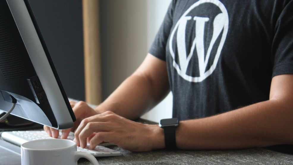 Eine Person schreibt auf einer Computertastatur. Sie trägt ein schwarzes T-Shirt mit dem WordPress-Logo und eine schwarze Armbanduhr. Im Vordergrund steht ein weißer Kaffeebecher.