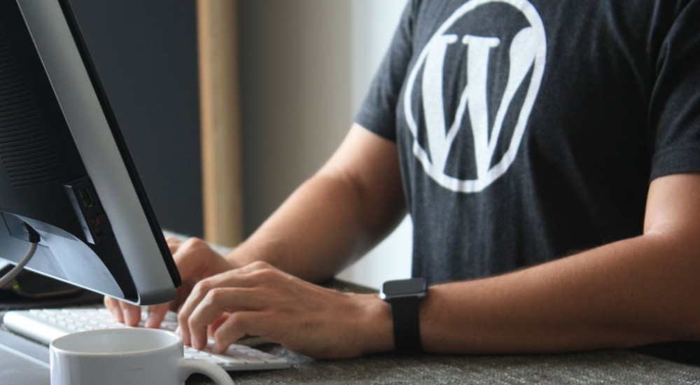 Eine Person schreibt auf einer Computertastatur. Sie trägt ein schwarzes T-Shirt mit dem WordPress-Logo und eine schwarze Armbanduhr. Im Vordergrund steht ein weißer Kaffeebecher.
