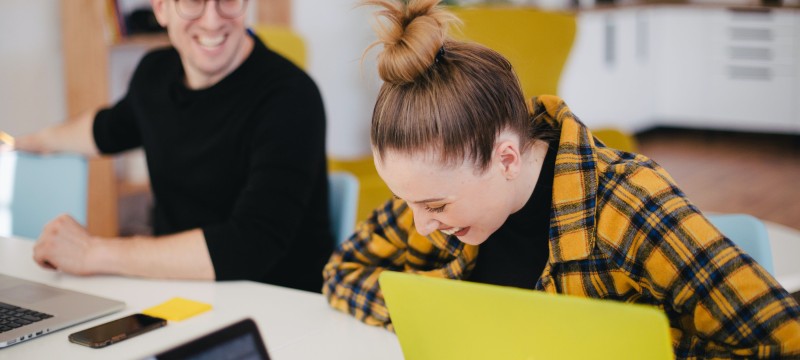 Zwei Kollegen sitzen am Schreibtisch und reden und lachen miteinander