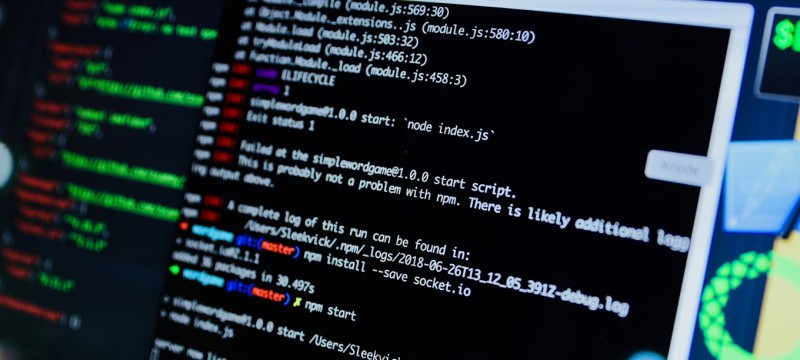Blick von schräg rechts auf einen Computerbildschirm. In einem Fenster ist Programmcode in weißer Schrift auf schwarzem Hintergrund zu sehen. Manche Code-Elemente sind durch unterschiedliche Schriftfarben hervorgehoben.