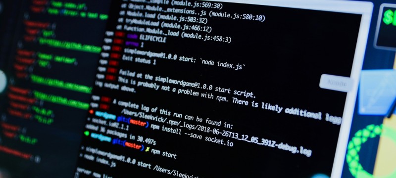 Blick von schräg rechts auf einen Computerbildschirm. In einem Fenster ist Programmcode in weißer Schrift auf schwarzem Hintergrund zu sehen. Manche Code-Elemente sind durch unterschiedliche Schriftfarben hervorgehoben.