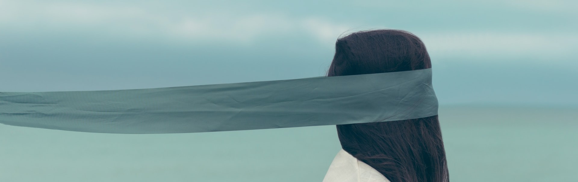 Eine Frau in hellem Oberteil mit langen, braunen Haaren ist von der Seite zu sehen. Ein schmales, blaues Tuch bedeckt ihre Augen und wird waagrecht nach hinten weggeweht. Im Hintergrund ist verschwommen eine Wasseroberfläche und bewölkter Himmel zu sehen.