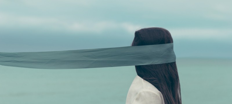 Eine Frau in hellem Oberteil mit langen, braunen Haaren ist von der Seite zu sehen. Ein schmales, blaues Tuch bedeckt ihre Augen und wird waagrecht nach hinten weggeweht. Im Hintergrund ist verschwommen eine Wasseroberfläche und bewölkter Himmel zu sehen.