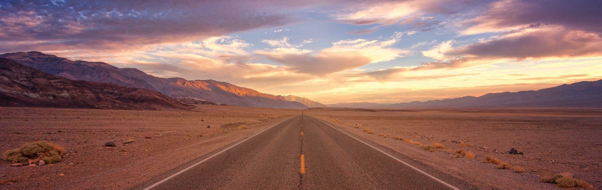 Eine asphaltierte Straße führt scheinbar endlos geradeaus. Die Umgebung ist eine Wüste, am Bildrand sind Hügel. Am Himmel sind Wolken, die von der untergegangenen Sonne in warme Farben getaucht werden.