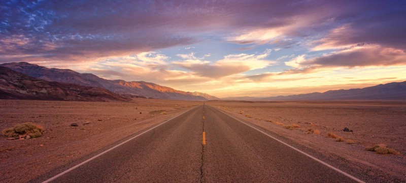 Eine asphaltierte Straße führt scheinbar endlos geradeaus. Die Umgebung ist eine Wüste, am Bildrand sind Hügel. Am Himmel sind Wolken, die von der untergegangenen Sonne in warme Farben getaucht werden.