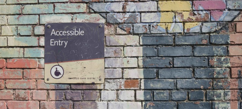 Auf einer bunt bemalten Mauer ist ein schwarz-gelbes Schild mit der Aufschrift "Accessible Entry" angebracht. Darunter ist das Symbol für einen Menschen im Rollstuhl abgebildet.