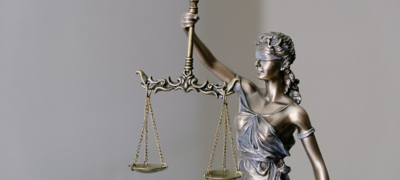 Eine Metallstatue der römischen Göttin Justitia vor hellem Hintergrund. Mit dem rechten Arm hält sie eine Waage hoch. Die beiden Waagschalen befinden sich auf gleicher Höhe. Die Augen der Göttin sind mit einem Tuch verbunden.