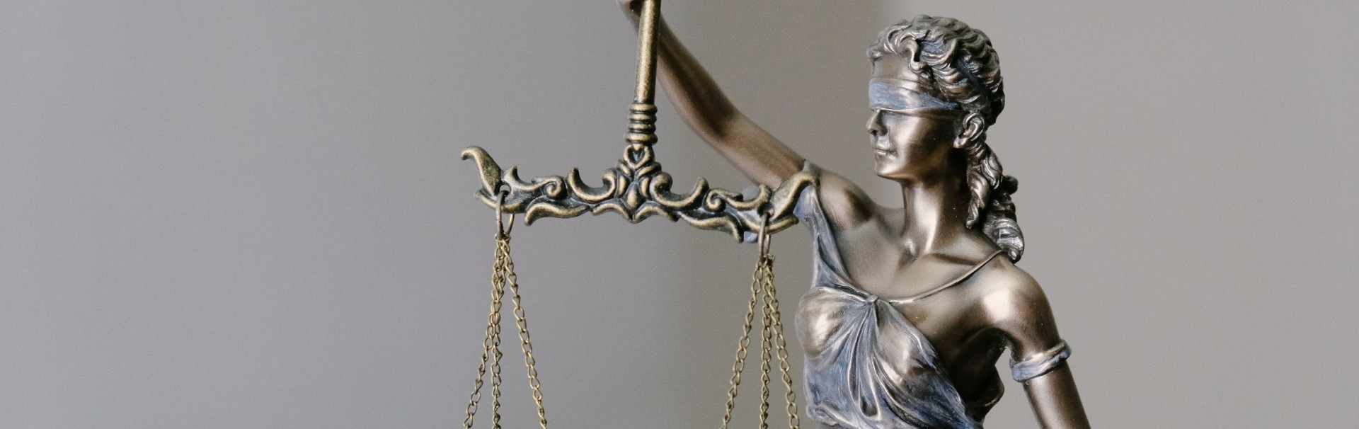 Eine Metallstatue der römischen Göttin Justitia vor hellem Hintergrund. Mit dem rechten Arm hält sie eine Waage hoch. Die beiden Waagschalen befinden sich auf gleicher Höhe. Die Augen der Göttin sind mit einem Tuch verbunden.