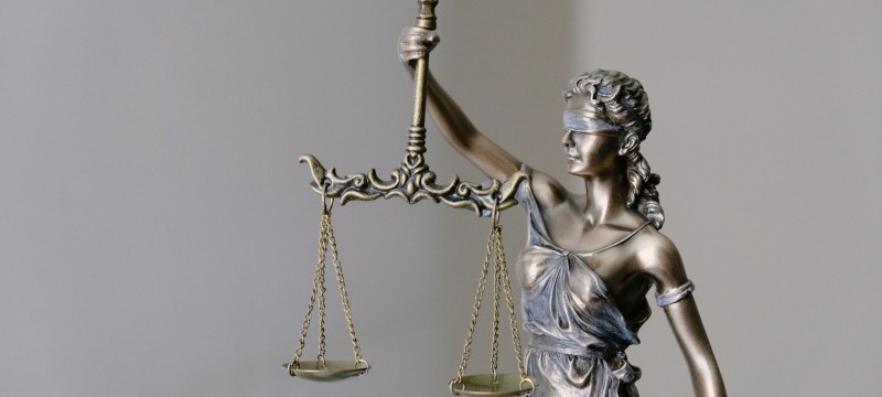 Eine Metallstatue der römischen Göttin Justitia vor hellem Hintergrund. Mit dem rechten Arm hält sie eine Waage hoch. Die beiden Waagschalen befinden sich auf gleicher Höhe. Die Augen der Göttin sind mit einem Tuch verbunden.