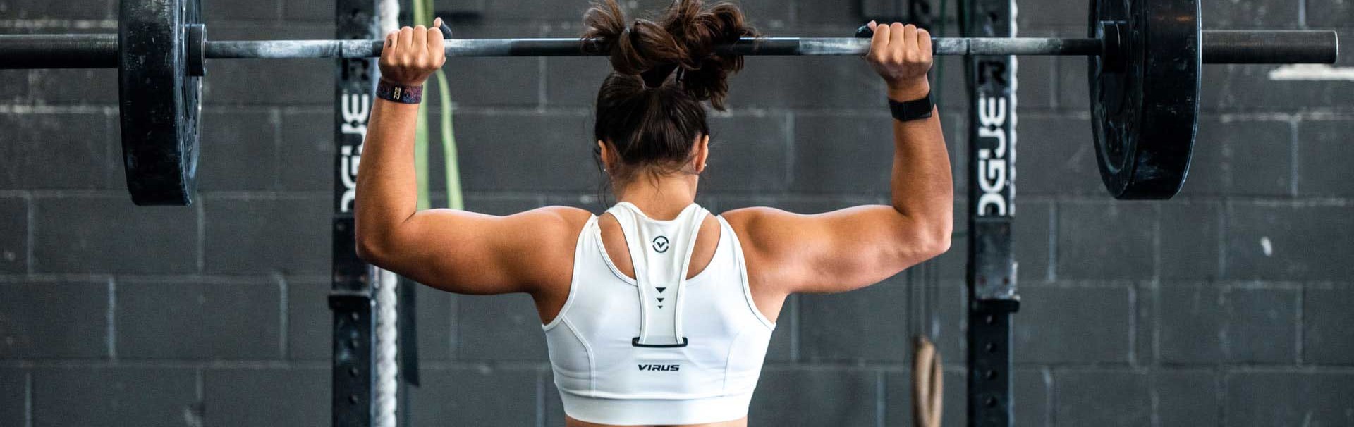 Es ist eine Frau von hinten beim Gewichtheben zu sehen. Sie trägt ein weißes Tanktop und stemmt die Langhantel über ihren Kopf.