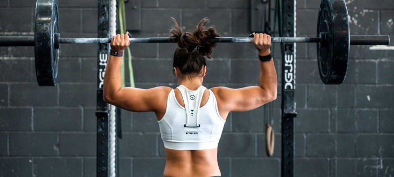 Es ist eine Frau von hinten beim Gewichtheben zu sehen. Sie trägt ein weißes Tanktop und stemmt die Langhantel über ihren Kopf.