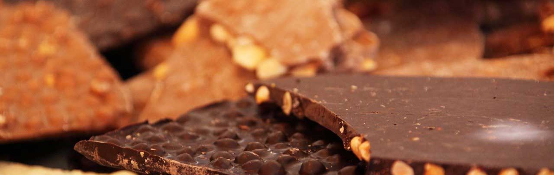 Verschiedene Sorten Schokolade liegen vermischt auf einem Haufen gestapelt. Die dünnen Tafeln sind teilweise gebrochen, sodass die Nussstücke im Inneren sichtbar sind