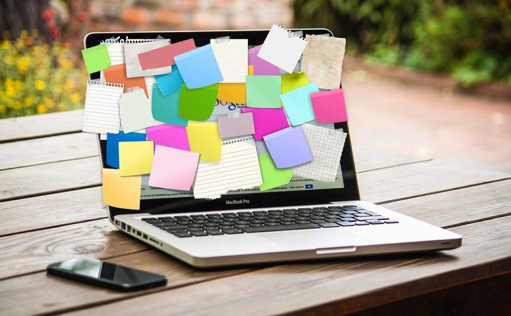 In einer Fotomontage kleben auf einem Laptop-Bildschirm so viele Haftnotizen, dass er ganz verdeckt ist. Der Laptop steht draußen auf einem Holztisch