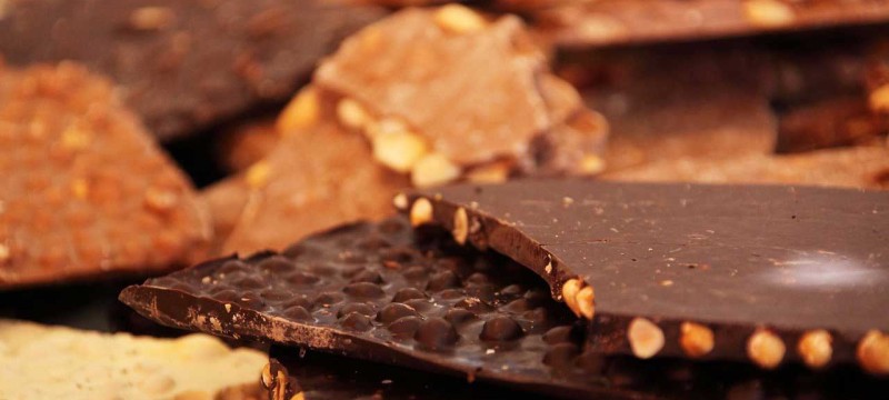 Verschiedene Sorten Schokolade liegen vermischt auf einem Haufen gestapelt. Die dünnen Tafeln sind teilweise gebrochen, sodass die Nussstücke im Inneren sichtbar sind