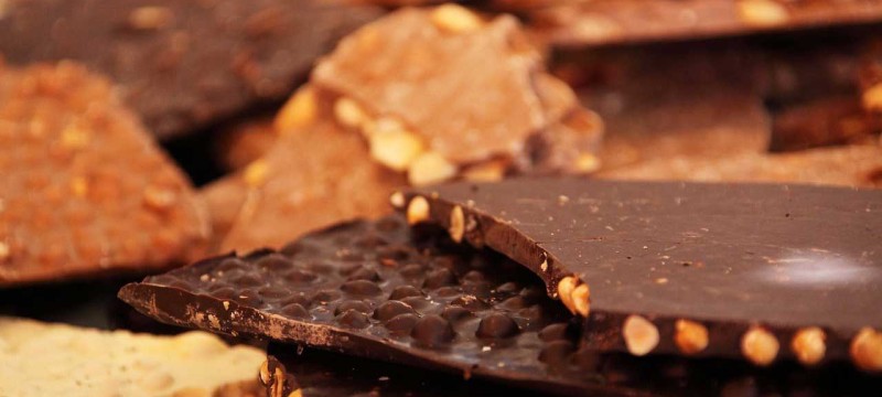 Verschiedene Sorten Schokolade liegen vermischt auf einem Haufen gestapelt. Die dünnen Tafeln sind teilweise gebrochen, sodass die Nussstücke im Inneren sichtbar sind