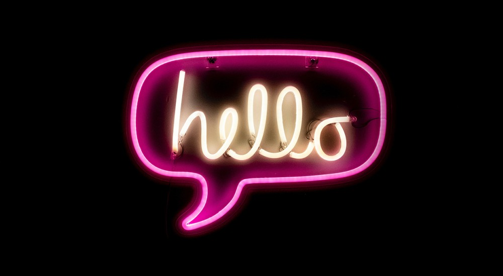 Vor einem schwarzen Hintergrund leuchtet eine pinkfarbene Sprechblase aus Neonröhren. Darin steht in weißer Schrift das Wort hello.