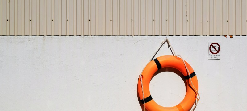 Ein orangefarbener Rettungsring hängt an einer hellgrauen Wand. Daneben hängt ein Verbotsschild mit der Aufschrift Nao mergulhar - No diving.
