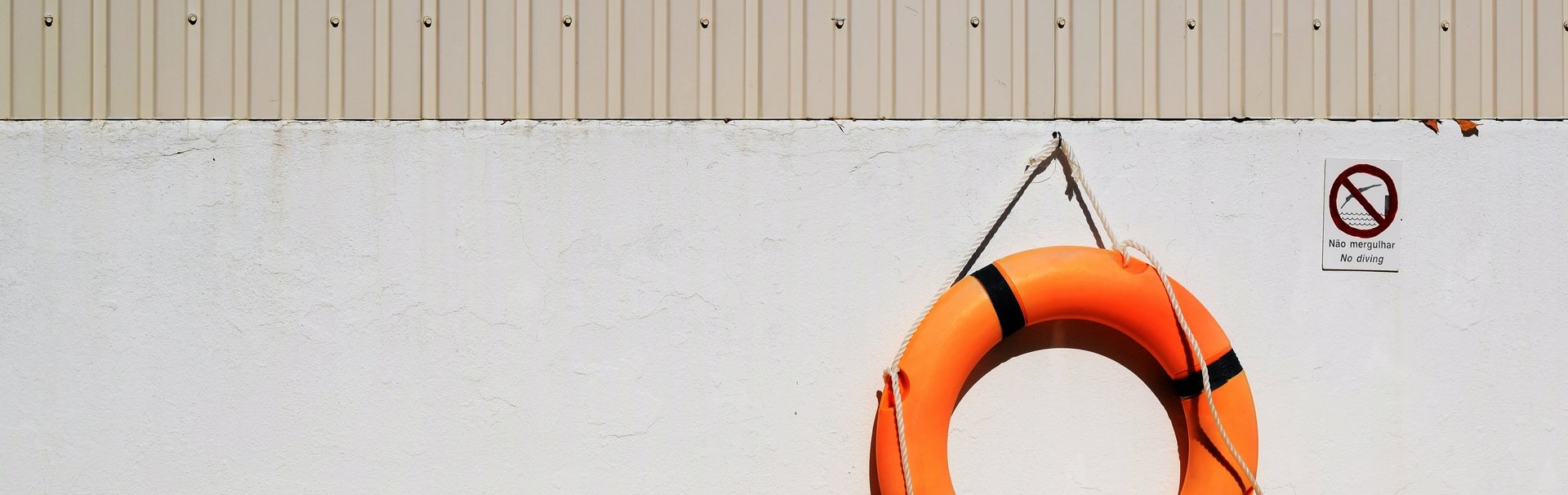 Ein orangefarbener Rettungsring hängt an einer hellgrauen Wand. Daneben hängt ein Verbotsschild mit der Aufschrift Nao mergulhar - No diving.