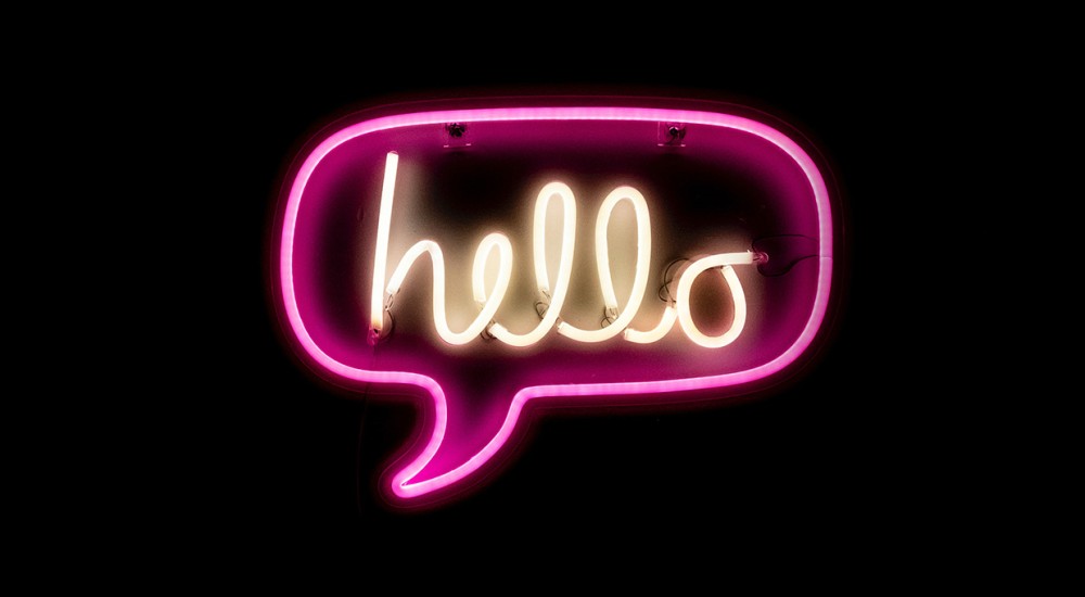 Vor einem schwarzen Hintergrund leuchtet eine pinkfarbene Sprechblase aus Neonröhren. Darin steht in weißer Schrift das Wort hello.