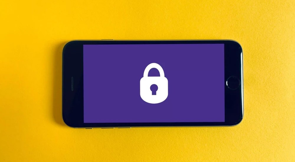 Auf einem gelben Untergrund liegt ein Smartphone. Auf seinem Bildschirm ist ein weißes Vorhängeschloss vor lila Hintergrund zu sehen.