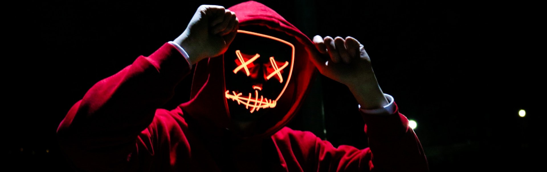 Vor einem dunklen Hintergrund steht eine Person im roten Kapuzenpulli und mit weißen Handschuhen. Die Person trägt eine Maske, die mit orangefarbenen Lichtern ein Gesicht wie auf einer Comic-Zeichnung darstellt. Die Augen sind zwei Kreuzchen.