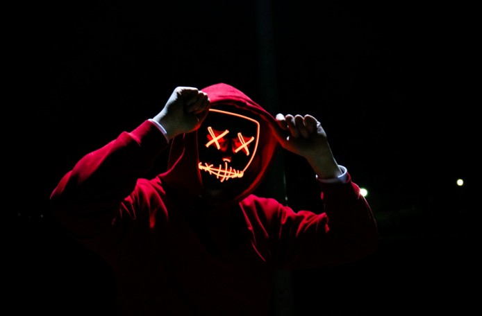 Vor einem dunklen Hintergrund steht eine Person im roten Kapuzenpulli und mit weißen Handschuhen. Die Person trägt eine Maske, die mit orangefarbenen Lichtern ein Gesicht wie auf einer Comic-Zeichnung darstellt. Die Augen sind zwei Kreuzchen.