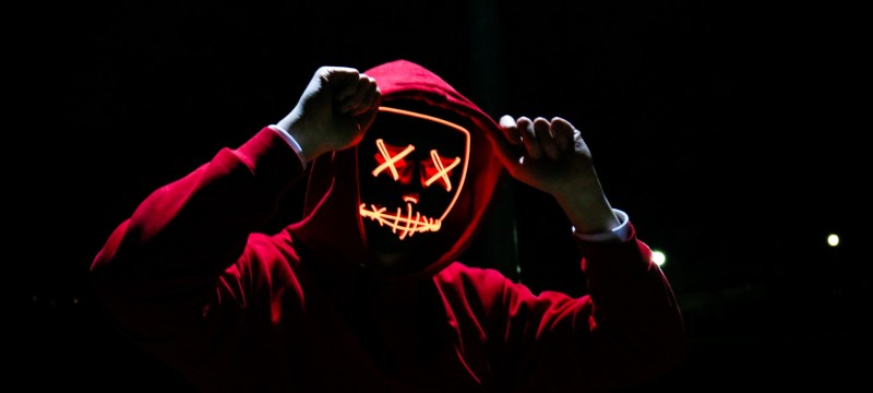 Vor einem dunklen Hintergrund steht eine Person im roten Kapuzenpulli und mit weißen Handschuhen. Die Person trägt eine Maske, die mit orangefarbenen Lichtern ein Gesicht wie auf einer Comic-Zeichnung darstellt. Die Augen sind zwei Kreuzchen.