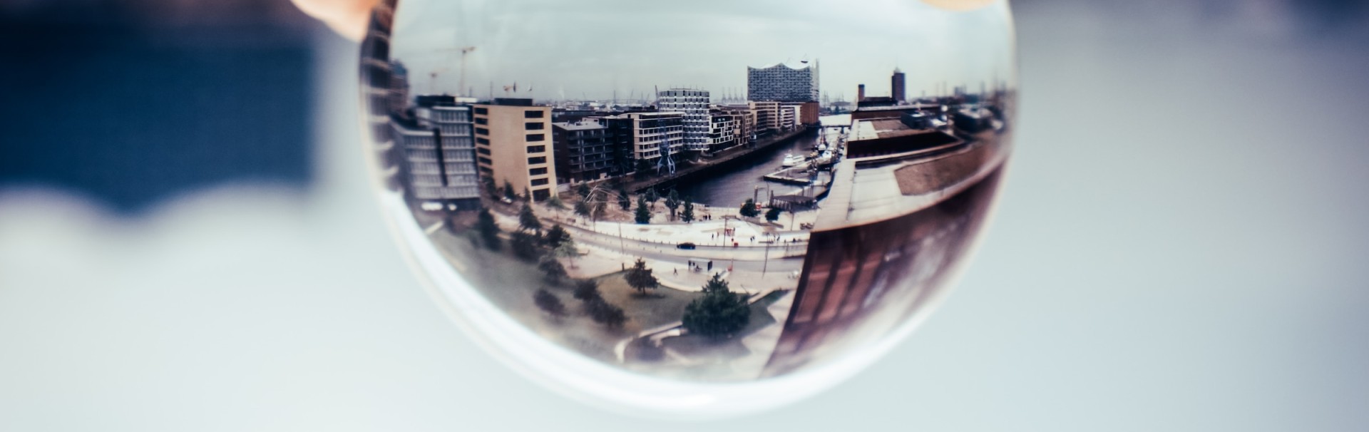 Eine städtische Landschaft, die durch eine Kristallkugel betrachtet wird, welche von einer Hand gehalten wird. Die Kugel zeigt eine umgedrehte, verzerrte Darstellung von modernen Gebäuden, einer Wasserstrasse und einem Platz mit Menschen. Die Umgebung ausserhalb der Kugel ist unscharf, was den Fokus auf die reflektierte Szene in der Kugel lenkt.