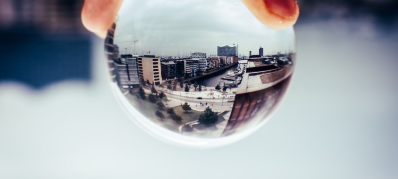 Eine städtische Landschaft, die durch eine Kristallkugel betrachtet wird, welche von einer Hand gehalten wird. Die Kugel zeigt eine umgedrehte, verzerrte Darstellung von modernen Gebäuden, einer Wasserstrasse und einem Platz mit Menschen. Die Umgebung ausserhalb der Kugel ist unscharf, was den Fokus auf die reflektierte Szene in der Kugel lenkt.