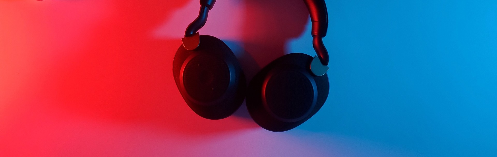 Ein Paar schwarze Kopfhörer hängt an einer weißen Wand. Die Ohrmuscheln sind zur Wand hin gedreht. Von links scheint rotes Licht, von rechts blaues Licht.