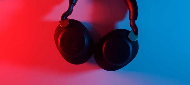 Ein Paar schwarze Kopfhörer hängt an einer weißen Wand. Die Ohrmuscheln sind zur Wand hin gedreht. Von links scheint rotes Licht, von rechts blaues Licht.