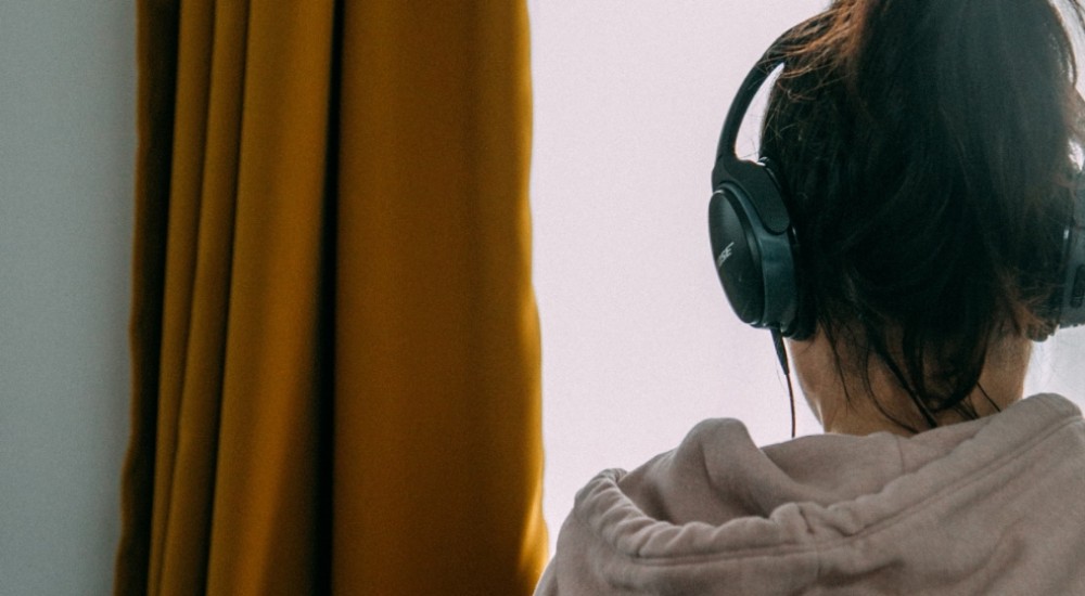 Blick von hinten auf eine Person, die vor einem hellen Fenster steht und nach draußen blickt. Der orangefarbene Vorhang ist nach links zur Seite geschoben. Die Person trägt einen hellen Kapuzenpulli und hat die braunen Haare zum Pferdeschwanz gebunden. Sie hat ein Paar große, schwarze Kopfhörer auf dem Kopf.
