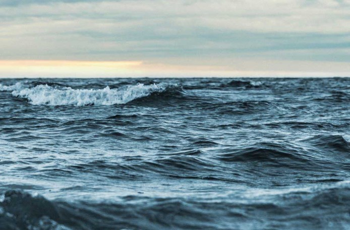 Unter einem grauen Himmel sieht man eine bewegte Meeresoberfläche, einzelne Wellen brechen.