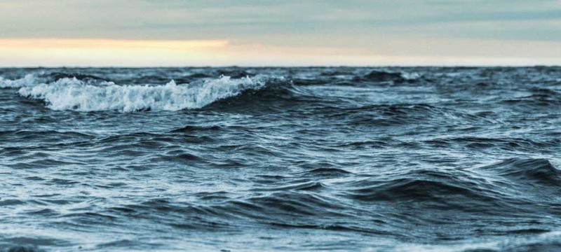 Unter einem grauen Himmel sieht man eine bewegte Meeresoberfläche, einzelne Wellen brechen.
