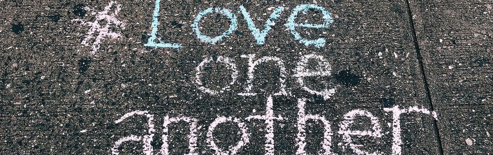 #Love one another steht mit bunter Kreide auf graue Gehwegplatten geschrieben - SCHOKOLADENSEITE.net übernimmt Verantwortung.